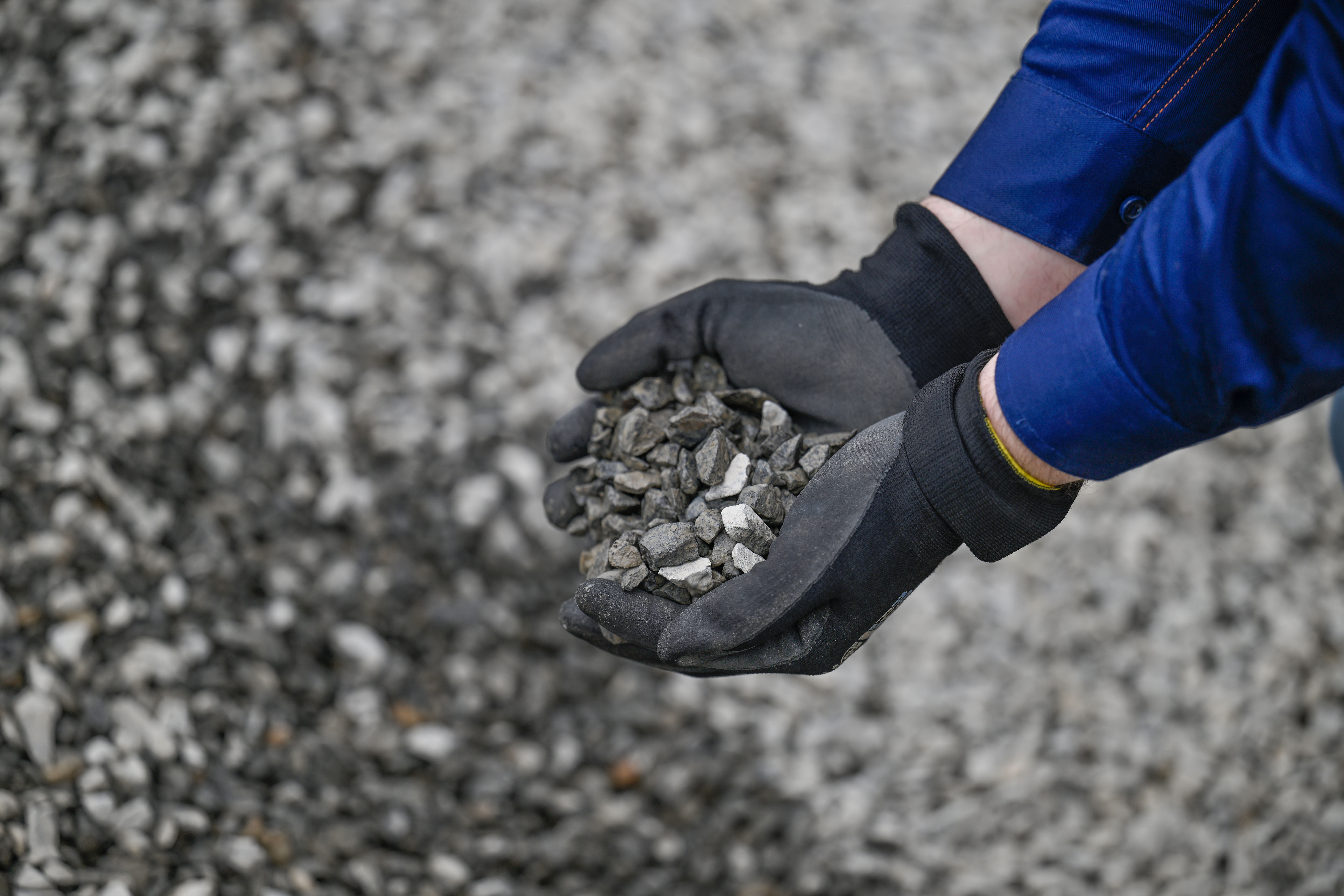 Gloved hands holding a small pile of gray gravel, with a larger gravel pile visible in the background.