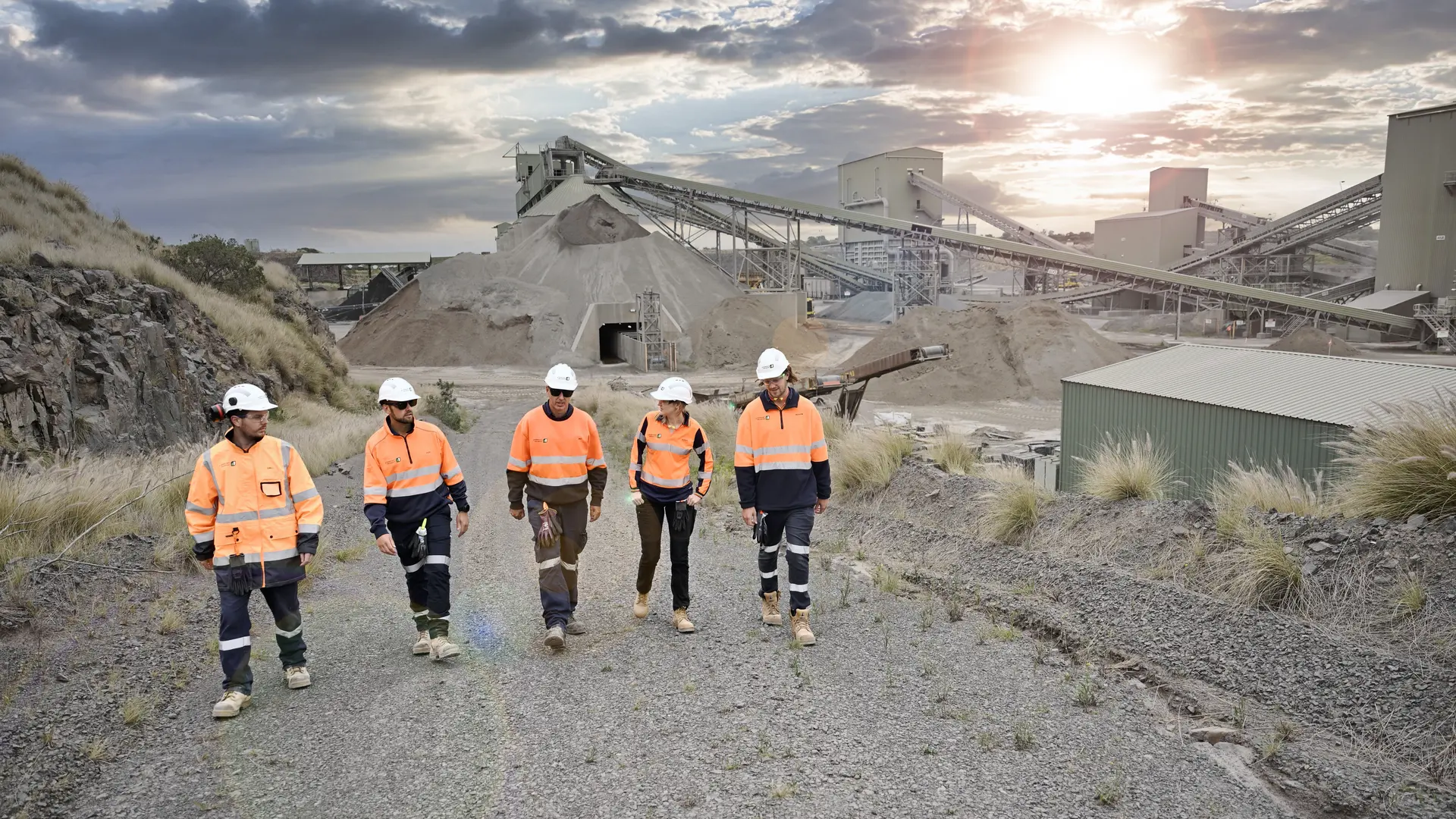 Heidelberg Materials employees wearing PPE and hard hats walking through a quarry site with processing facilities and aggregate stockpiles in the background.