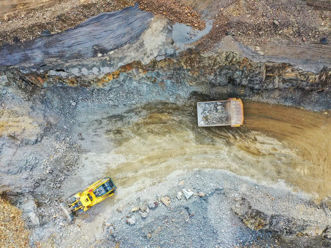 Aerial view of quarry with a yellow excavator on the bottom left and a large dump truck on the top right. The excavation area shows exposed layers of earth, rocks, and dirt. The dump truck appears to be partially loaded, positioned on a dirt pathway within the pit.