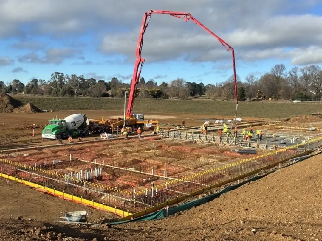 Concrete pump and Heidelberg Materials agitator truck pouring concrete into reinforced foundations at a large commercial construction site, with workers preparing the concrete slab.