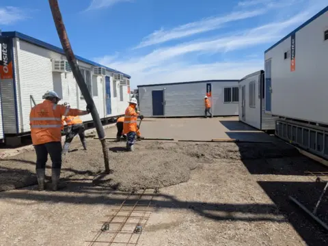 Construction workers complete a concrete pour using calcined clay concrete supplied by Heidelberg Materials Australia at a temporary site compound with modular buildings and reinforced slab preparation.